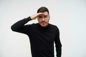 Portrait of young handsome dark haired man in eyewear squinting his eyes and keeping raised hand on forehead while trying to see something far away, posing over white background photo