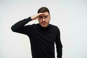 retrato de joven hermoso oscuro peludo hombre en gafas bizco su ojos y acuerdo elevado mano en frente mientras molesto a ver alguna cosa lejos lejos, posando terminado blanco antecedentes foto