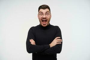 Excited young lovely bearded brunette male crossing arms on his chest and looking amazedly at camera with wide eyes and mouth opened, standing over white background photo