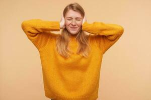 interior foto de encantador joven largo peludo dama con casual peinado sonriente ligeramente con cerrado ojos y participación elevado palmas en su orejas, posando terminado beige antecedentes