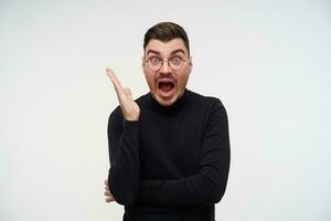 Shocked young short haired bearded brunette guy keeping his palm raised while looking surprisedly at camera, wearing elegant wear while posing over white background photo