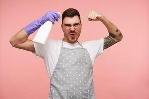 Joyful young tattooed brunette male with short haircut raising hands and looking self-confidently at camera, wearing basic white t-shirt and apron while standing over pink background photo