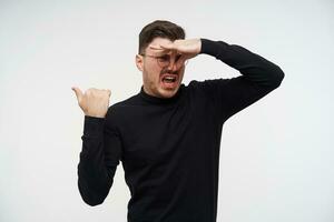 Indoor photo of displeased young dark haired guy in eyewear closing his nose while showing disgust and pointing aside with raised hand, isolated over white background