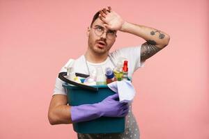 Tired young bearded dark haired guy in eyeglasses wiping sweat with raised hand while being tired after spring cleaning, standing over pink background in working wear photo