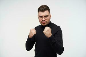 Serious young dark haired man with short haircut raising hands in protective gesture while looking severely at camera, standing over white background photo