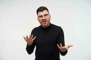 Puzzled young handsome unshaved brunette man raising confusedly hands and grimacing his face while looking at camera, standing over white background photo