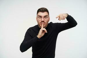 Portrait of young angry short haired brunette male pointing crossly at camera with forefinger while showing hush gesture, isolated over white background photo