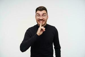 Excited young short haired bearded brunette man raising hand with hush gesture and looking excitedly at camera, wearing black sweater while standing over white background photo