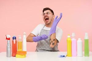 Indoor photo of tattooed dark haired male in working clothes screaming with closed eyes while putting on purple rubber gloves, isolated over pink background