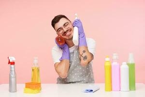 Pleased young good looking brunette man in eyeglasses keeping spray bottles with household chemicals and smiling happily with closed eyes, isolated over pink background photo