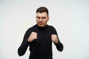 Severe young handsome bearded brunette guy in glasses keeping fists raised and frowning eyebrows while looking at camera, isolated over white background photo