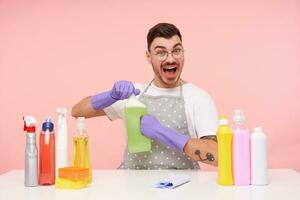 Agitated young lovely short haired brunette male being excited while opening bottle with household chemicals, wearing uniform and rubber gloves while sitting over pink background photo