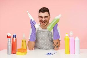 Indoor shot of cheerful young pretty short haired brunette man holding bottles of household chemicals and smiling happily while sitting over pink background photo