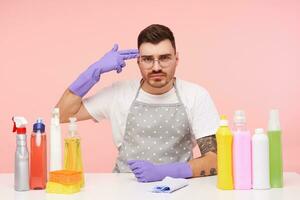 Displeased young short haired brunette guy with beard raising hand in rubber glove with gun gesture and looking at camera with folded lips, isolated over pink background photo