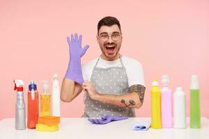 Overjoyed young bearded dark haired male with short haircut looking joyfully at camera with wide mouth opened and keeping his hand raised, posing over pink background photo