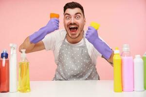 Agitated young pretty short haired brunette male looking excitedly at camera with wide eyes and mouth opened and raising hands with cleaning sponges, isolated over pink background photo