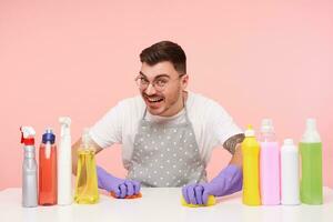 Joyful young lovely short haired brunette male in rubber wearing rubber gloves while washing table with sponges and smiling widely at camera, sitting over pink background photo
