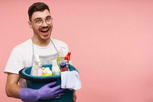 Joyful young pretty short haired brunette guy in glasses being in high spirit while finishing spring cleaning and smiling happily at camera, isolated over pink background photo