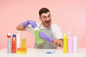 alegre joven bonito corto peludo morena chico en lentes vestido en trabajando ropa y guantes sonriente felizmente mientras apertura botella con casa quimicos, aislado terminado rosado antecedentes foto