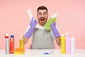 Overjoyed young attractive unshaved brunette man in glasses looking excitedly at camera and keeping household chemicals in raised hands, isolated over pink background photo