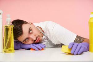 Weary young unshaved brunette man being tired after spring cleaning and leaning head on hand in rubber glove, dressed in uniform while sitting over pink background photo