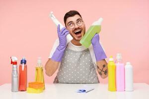 alegre joven corto peludo sin afeitar morena chico vestido en trabajando ropa y caucho guantes mirando felizmente hacia arriba mientras sentado terminado rosado antecedentes con detergentes foto