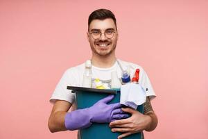 Cheerful young attractive brunette male with short haircut smiling widely at camera while keeping busket with household chemicals in raised hands, isolated over pink background photo