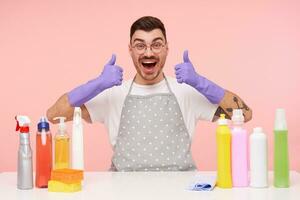 Indoor photo of overjoyed young lovely dark haired man with beard raising hands with thumbs and smiling cheerfully at camera, posing over pink background with detergents