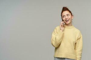 Nice looking woman, beautiful redhead girl with hair bun. Wearing yellow sweater and talking on a phone. Watching with a smile to the left at copy space, isolated over grey background photo