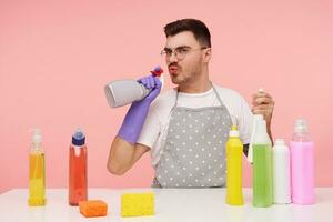 Studio photo of young funny brown haired male in glasses dressed in working clothes and rubber gloves blowing on spray bottle in his hand while imitating gun with it, isolated over pink background