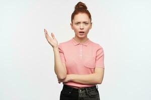 Unhappy lady, frowning woman with ginger hair bun. Wearing pink t-shirt and black jeans. Raise her hand in disagreement. Watching at the camera, isolated over white background. photo