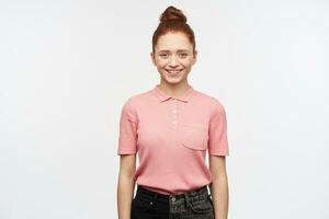 Nice looking woman, beautiful redhead girl with hair bun. Wearing pink t-shirt and black jeans. Have a cheerful mood and broadly smiling. Watching at the camera, isolated over white background. photo