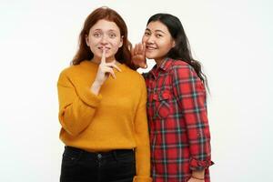 People and lifestyle concept. Two hipster friends. Girl whisper to her friend, showing silence sign. Wearing yellow sweater and checkered shirt. Watching at the camera, isolated over white background photo