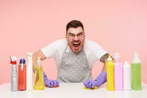 Annoyed young dark haired unshaved man in glasses being tired after cleaning house and screaming angrily while washing table, isolated over pink background photo