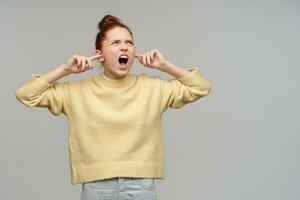 Unhappy, screaming girl with ginger hair gathered in a bun. Wearing pastel yellow sweater and jeans. Close her ears and yells. Watching to the right at copy space, isolated over grey background photo