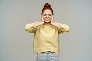 Nice looking woman, beautiful girl with ginger hair gathered in a bun. Wearing pastel yellow sweater and jeans. Cover her ears and squints. Do not want to hear. Stand isolated over grey background photo