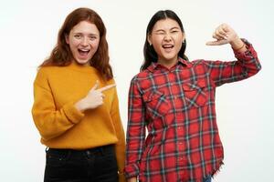 Portrait of cheerful asian and caucasian friends. Wearing casual outfit. Happy girl pointing on her friend who winks and pointing on her self. Watching at the camera, isolated over white background photo
