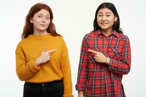 Portrait of asian and caucasian girls. Hipster friends pointing, blame each other. Wearing yellow sweater and checkered shirt. People concept. Watching at the camera, isolated over white background photo