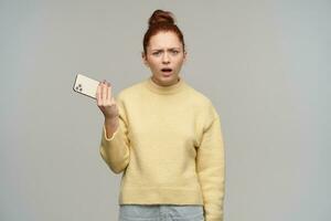 Confused lady, disconnected woman with ginger hair bun. Wearing yellow sweater and holding a smartphone. Frowning her face, bothered. Watching at the camera, isolated over grey background photo