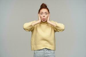 Teenage girl, funny looking woman with ginger hair gathered in a bun. Wearing pastel yellow sweater and jeans. Cover her ears and showing tongue. Watching at the camera isolated over grey background photo