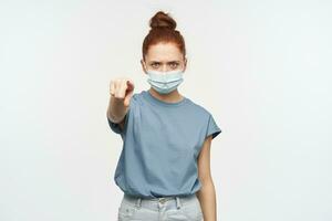 Portrait of frowning, serious redhead girl with hair gathered in a bun. Wearing blue t-shirt, jeans and protective face mask. Pointing at you. Watching at the camera isolated over white background photo