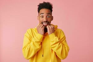 Scared young african american guy in yellow hoodie, with horror looking into the camera with wide open eyes and clenches teeth, bites finger nails, stands over pink background. photo