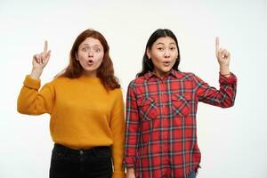 Two shocked, young women friends. Wearing yellow sweater and checkered shirt. People concept. Watching amazed at the camera and pointing up at copy space, isolated over white background photo