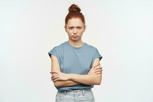 Teenage girl, unhappy looking woman with ginger hair gathered in a bun. Wearing blue t-shirt and jeans. Keep arms crossed, pout lip and frown. Watching at the camera isolated over white background photo