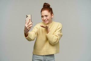 Cute lady, pretty woman with ginger hair bun. Wearing yellow sweater and holding a smartphone. Making selfie, have video call. Sending air kiss, smile. Stand isolated over grey background photo