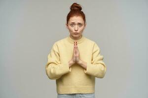 Sad woman with ginger hair gathered in a bun. Wearing pastel yellow sweater and jeans. Keep her palms together in prayer. Ask for something. Watching at the camera isolated over grey background photo