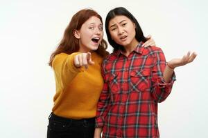 Two young women friends. Girl showing to her friend something, but another one can not see. Wearing yellow sweater and checkered shirt. Watching at the camera, isolated over white background photo