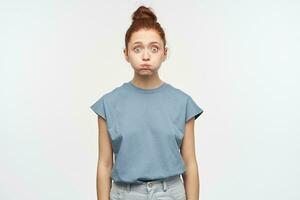 Surprised teenage girl, funny looking woman with ginger hair gathered in a bun. Wearing blue t-shirt and jeans. Puffed out her cheeks. Watching at the camera isolated over white background photo