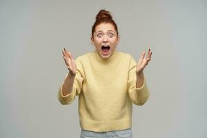 Horrified looking woman with ginger hair gathered in a bun. Wearing pastel yellow sweater and jeans. Screams in fear, terrified by what she see. Watching at the camera isolated over grey background photo