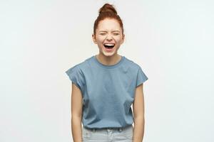 Funny woman, beautiful girl with ginger hair gathered in a bun. Wearing blue t-shirt and jeans. Laughing with closed eyes, hear hilarious joke. Emotion concept. Stand isolated over white background photo